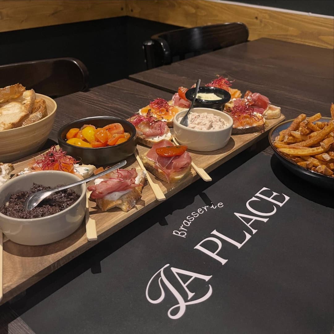 Une planche à partager sur une table de la brasserie : tartines garnies de jambon cru et de saumon, tartinades, tomates confites et frites, sur le set de table au logo La Place.